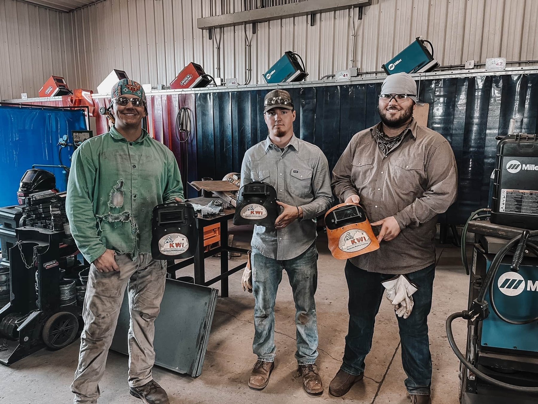 Three KWI students on the shop floor holding their KWI-branded welding hoods